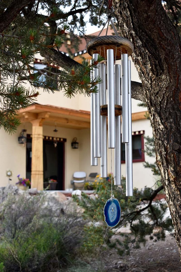 Metal wind chimes with a blue agate stone hanging from a tree near a house porch.
