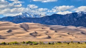 Sand dunes with green vegetation in front and snow-capped mountains under a partly cloudy sky.